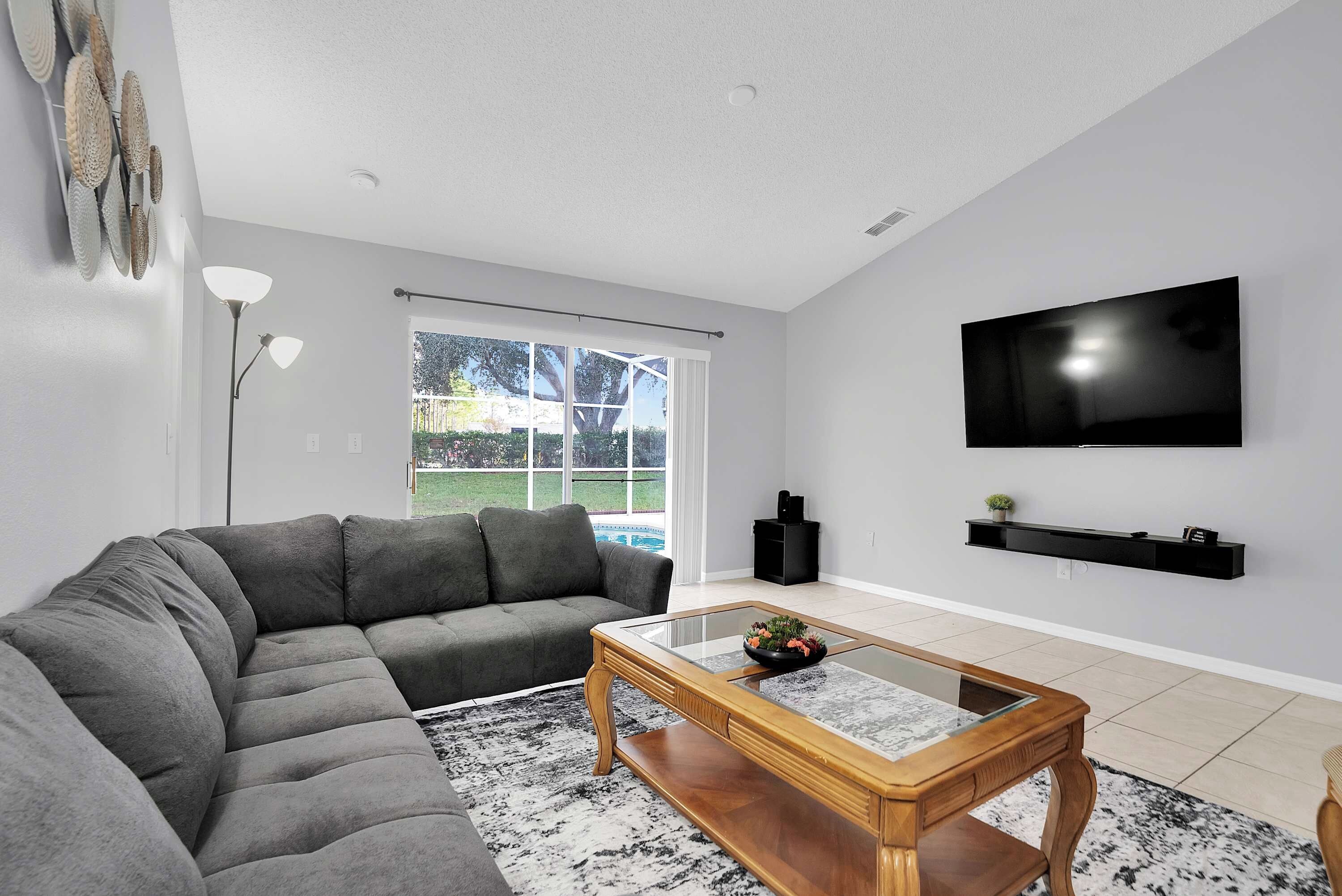 A living room with a sectional sofa, a coffee table, a flat-screen TV, and a window with a view of a swimming pool.