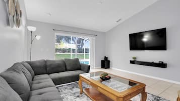 A living room with a sectional sofa, a coffee table, a flat-screen TV, and a window with a view of a swimming pool.