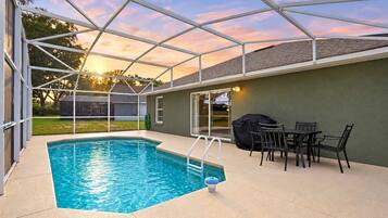 A covered outdoor swimming pool with a patio area, a barbecue grill, and a table with chairs.