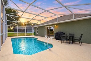 A covered outdoor swimming pool with a patio area, a barbecue grill, and a table with chairs.