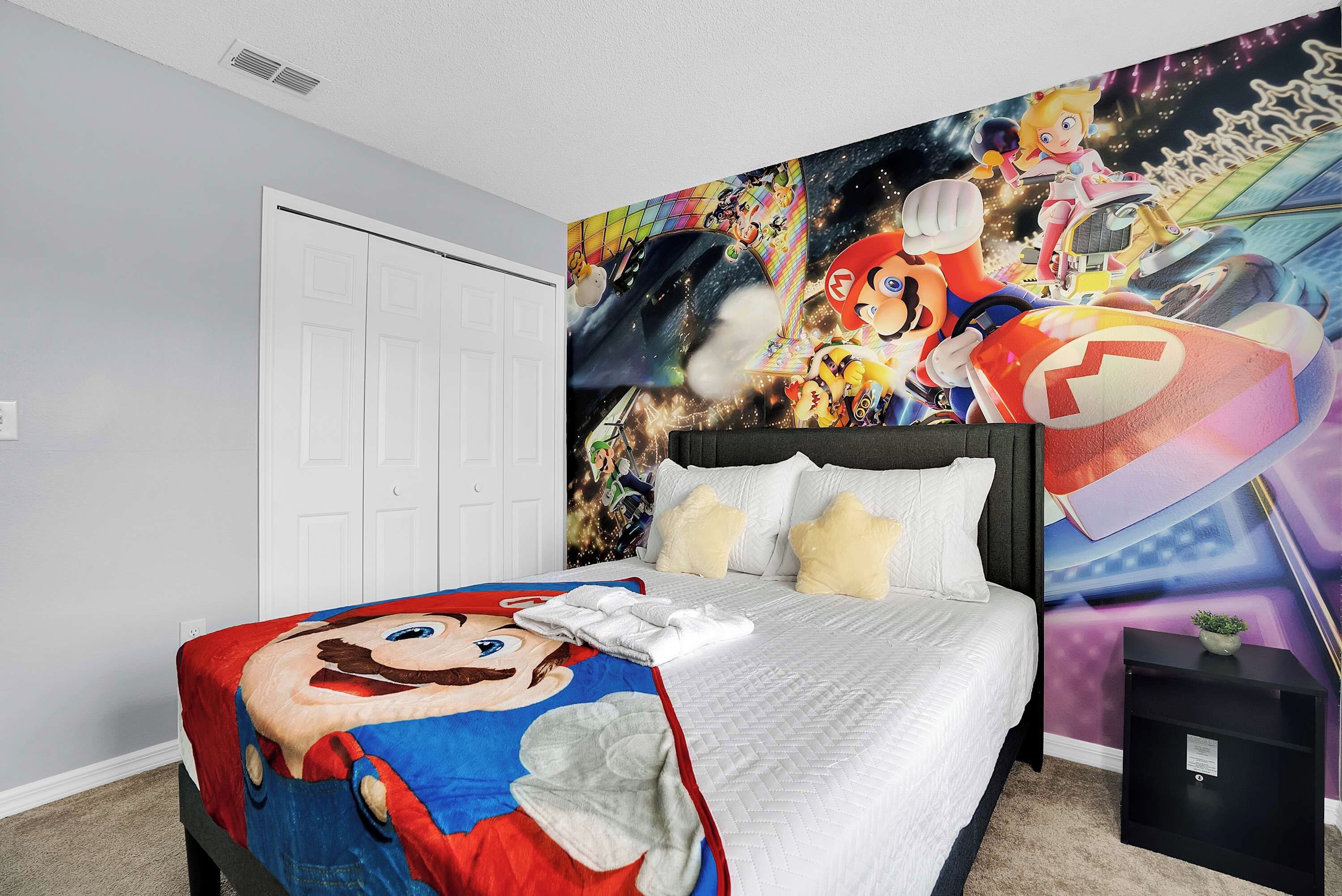 Een slaapkamer met een bed, een Mario-thema beddeken, een muurschildering met verschillende videospel personages en een witte deur.