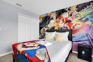 Een slaapkamer met een bed, een Mario-thema beddeken, een muurschildering met verschillende videospel personages en een witte deur.