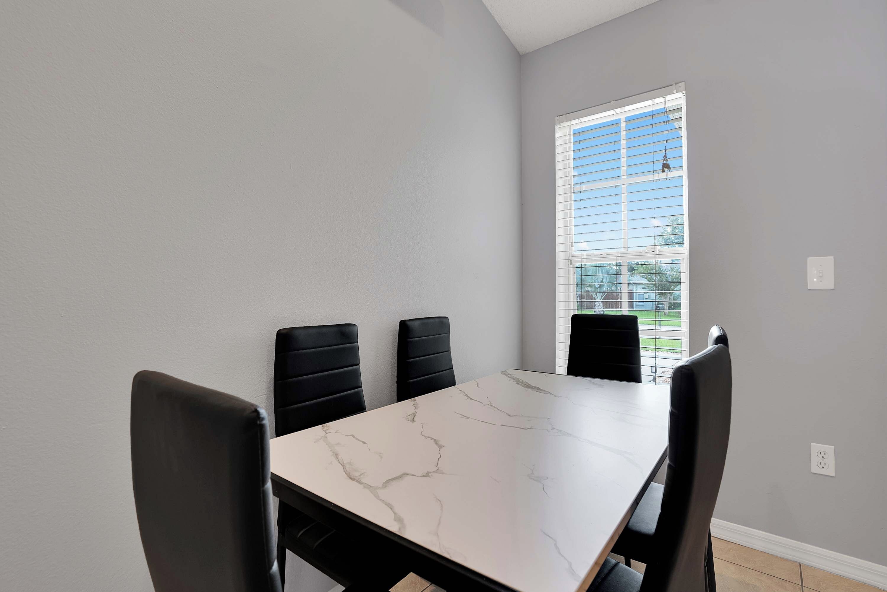 Un coin repas avec une table en marbre et des chaises noires, une fenêtre avec des stores et un mur de couleur gris clair.