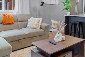 A modern living room with a grey sofa, a wooden coffee table, and a kitchen area in the background. - Upscale 2BR/2BA Apt | Mins to NAIT, MacEwan & DT - Apartment (Edmonton)