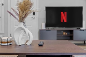 A living room with a television displaying the Netflix logo, a remote control, and a decorative vase with dried plants. - Upscale 2BR/2BA Apt | Mins to NAIT, MacEwan & DT - Apartment (Edmonton)