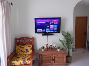 Ein Wohnzimmer mit einem an der Wand befestigten Flachbildfernseher, einem Holzschrank mit Dekorationen, einem stuhl mit Blumenmuster und einer Topfpflanze.