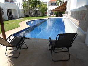 Ein Swimmingpool mit zwei Liegestühlen, umgeben von einem weißen Gebäude mit einem Kachel­dach, und Palmen im Hintergrund.