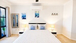 A bedroom with a bed, two bedside tables with lamps, a window with curtains, an air conditioning unit, and wall art. - **Between Two Blues – Beachfront Bliss in Yeppoon** (Yeppoon)