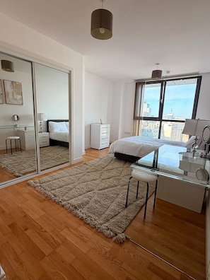 Ein modernes Schlafzimmer mit großem Spiegel, einem Bett, einem Schreibtisch und Blick auf die Stadt durch ein großes Fenster.