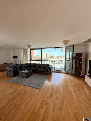 A modern living room with a large sofa, a coffee table, and a view of a cityscape through large windows. - Luxurious Mailbox Penthouse 3 Bedrooms 3 Bathrooms (Birmingham)