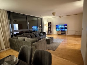 A modern living room with a large flat-screen TV, a sectional sofa, and a coffee table. - Luxurious Mailbox Penthouse 3 Bedrooms 3 Bathrooms (Birmingham)
