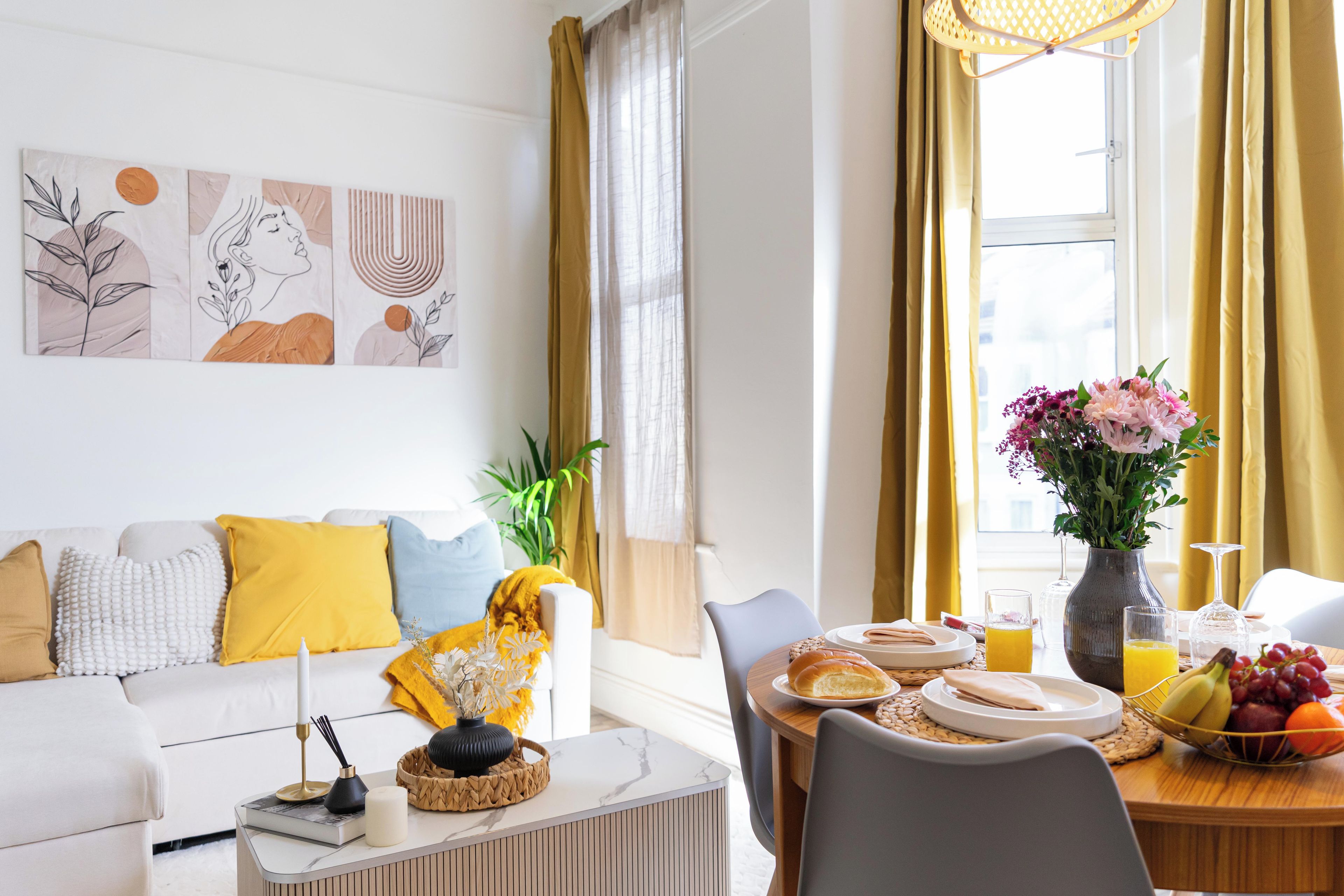 A modern living room with a sofa, a dining table set with breakfast items, and a large window with curtains.