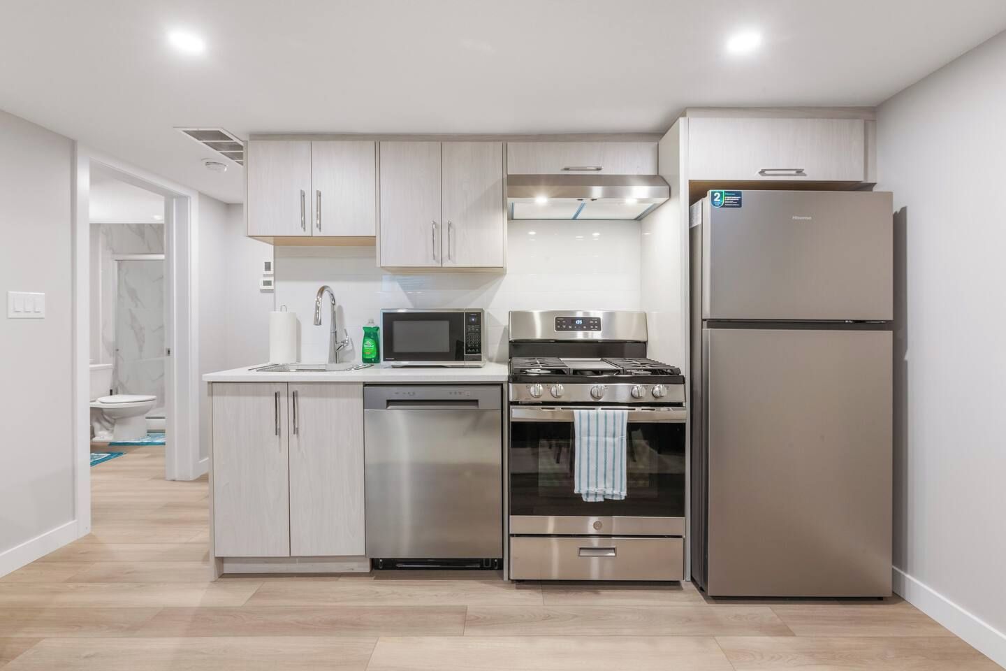 Una cucina moderna con elettrodomestici in acciaio inossidabile, mobili chiari in legno e un bagno in vista sullo sfondo.