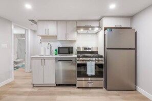 A modern kitchen with stainless steel appliances, light wood cabinetry, and a visible bathroom in the background.