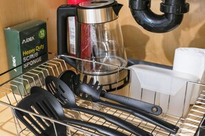 Una zona de cocina con una estructura metálica que contiene utensilios de cocina negros, un hervidor de acero inoxidable, una caja de esponja limpiadora AIDE y un recipiente blanco.