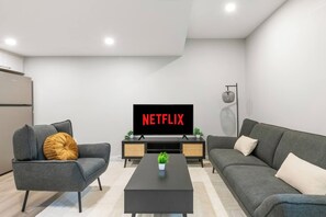 A modern living room with a flat-screen TV displaying 'NETFLIX,' a grey sofa, a grey armchair, a black coffee table, and a small plant. - Stylish 1BR Guest Suite | Near UofA, Whyte & DT - Double Room (Edmonton)