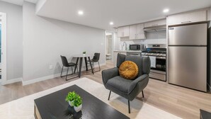 Une cuisine moderne équipée d’appareils électroménagers en acier inoxydable, un coin repas avec une table ronde et des chaises, et un fauteuil gris agrémenté d’un coussin décoratif.