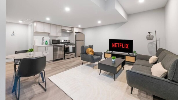Un salon moderne doté d’un téléviseur à écran plat affichant le logo « NETFLIX », d’un coin repas avec une table en verre et d’une cuisine équipée d’appareils électroménagers en acier inoxydable.