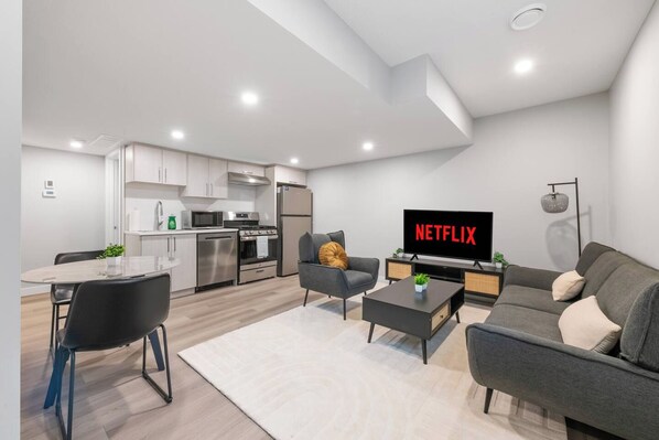 A modern living room with a flat-screen TV displaying 'NETFLIX,' a dining area with a glass table, and a kitchen with stainless steel appliances. - Stylish 1BR Guest Suite | Near UofA, Whyte & DT - Double Room (Edmonton)