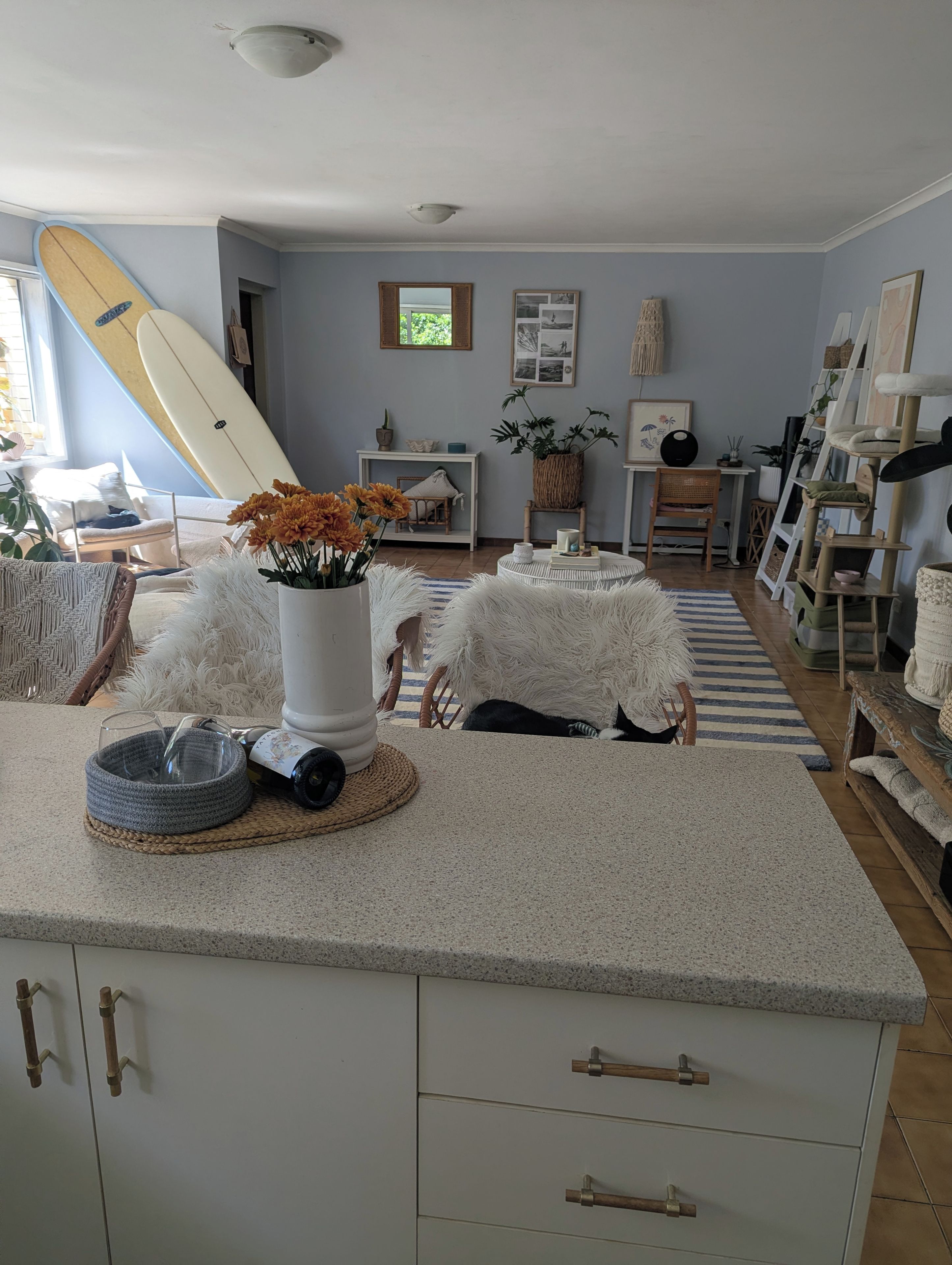 A living room with a surfboard leaning against the wall, a round coffee table with a vase of flowers, and a cat climbing on the couch.