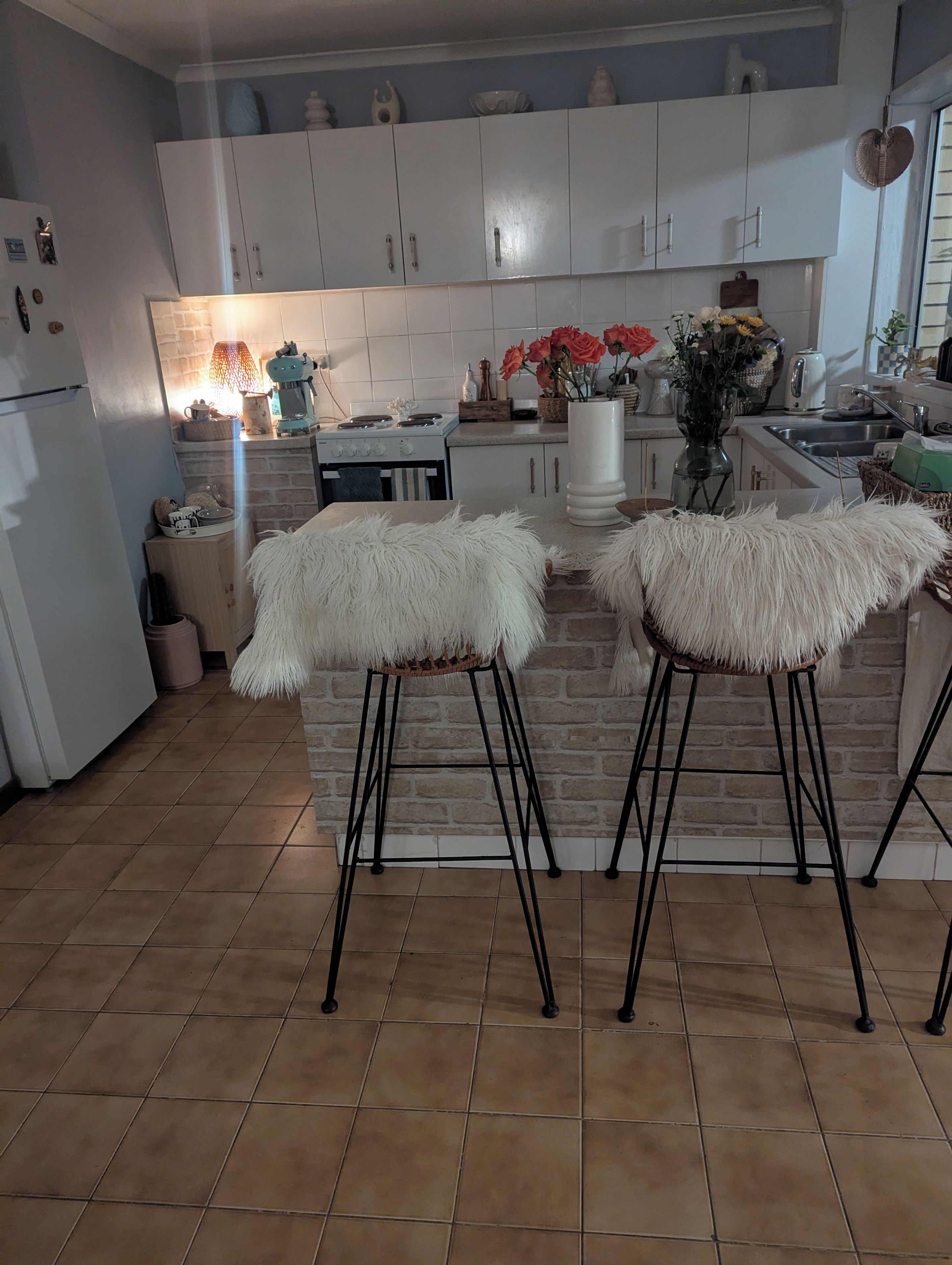 A kitchen with white cabinets, a brick backsplash, two white furry stools, and a variety of kitchen appliances and decorations.