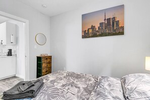 A bedroom with a bed, a dresser, a painting of a cityscape, and a bathroom visible through an open door. - Chic Getaway | 1BR Suite w/ Parking & AC (Windsor)