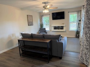 Ein Wohnzimmer mit einer Couch, einem Couchtisch, einem Deckenventilator, einem an der Wand montierten Fernseher und einem Kamin.