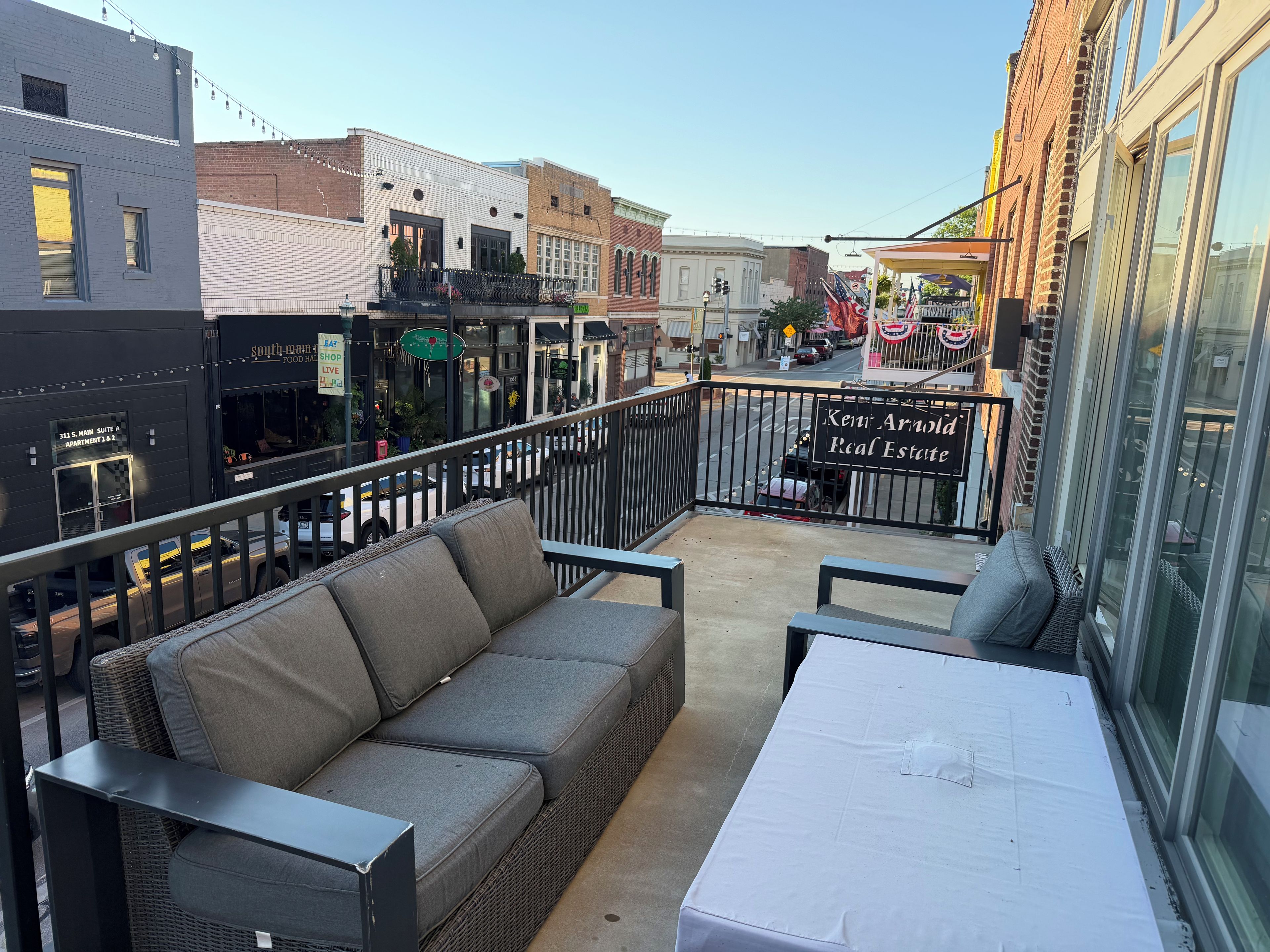 A balcony with a grey sofa and a table, overlooking a street with buildings and a sign for a real estate office.