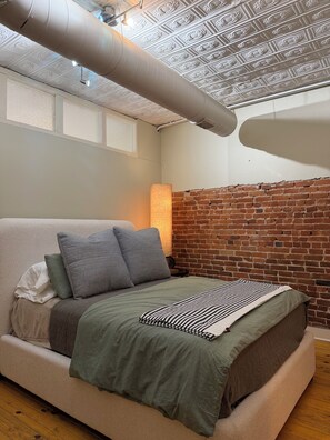 A modern bedroom with a brick wall, a bed with a green blanket, and a lamp.