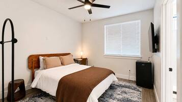A bedroom with a bed, a ceiling fan, a bedside table, a lamp, a TV, and a window with blinds.