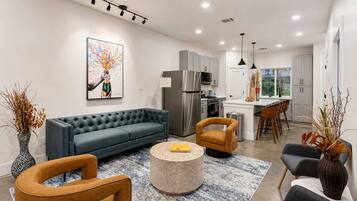 A modern living room with a grey sofa, round coffee table, and two armchairs. A kitchen with stainless steel appliances and a dining area with a bar stool is visible in the background.