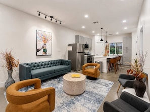 A modern living room with a grey sofa, round coffee table, and two armchairs. A kitchen with stainless steel appliances and a dining area with a bar stool is visible in the background. - Bishop Arts 6BD 6BA Townhouse Parking Sleeps 12 (Dallas)