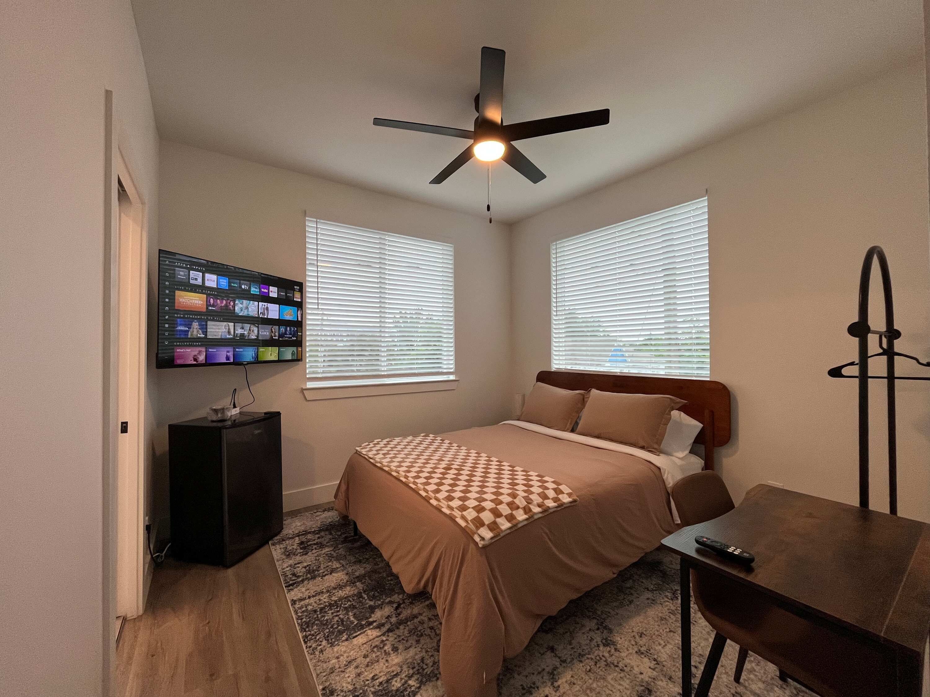 A bedroom with a bed, a desk, a television mounted on the wall, and a ceiling fan.