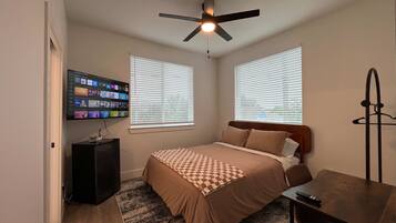 A bedroom with a bed, a desk, a television mounted on the wall, and a ceiling fan.