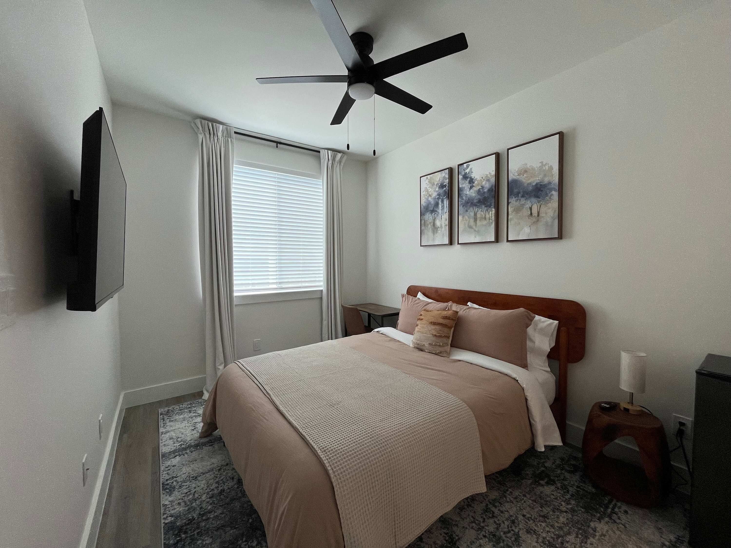 A bedroom with a bed, a ceiling fan, a window with curtains, and three framed pictures on the wall.