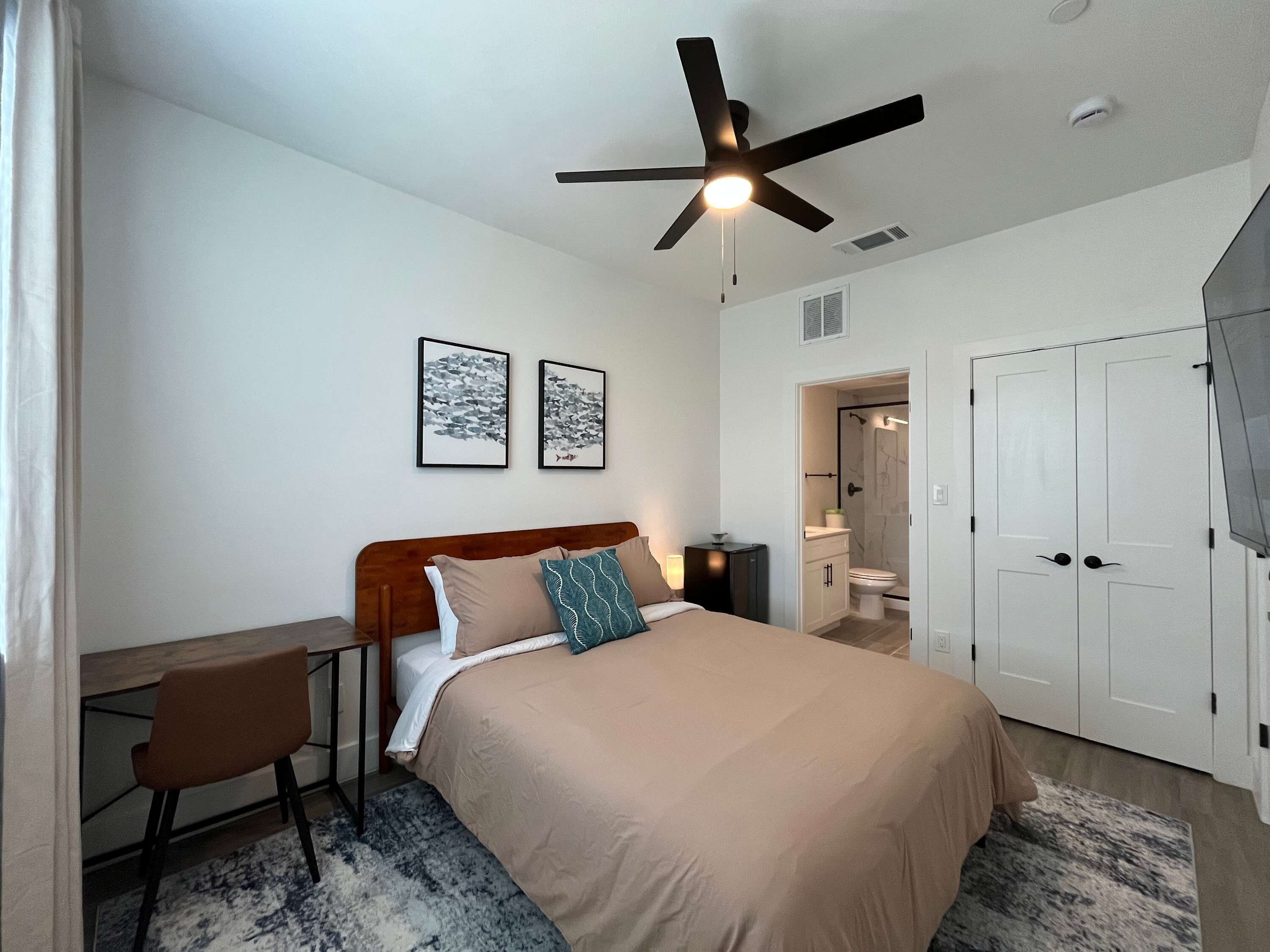 A bedroom with a bed, desk, chair, ceiling fan, artwork, and a bathroom visible through an open door.