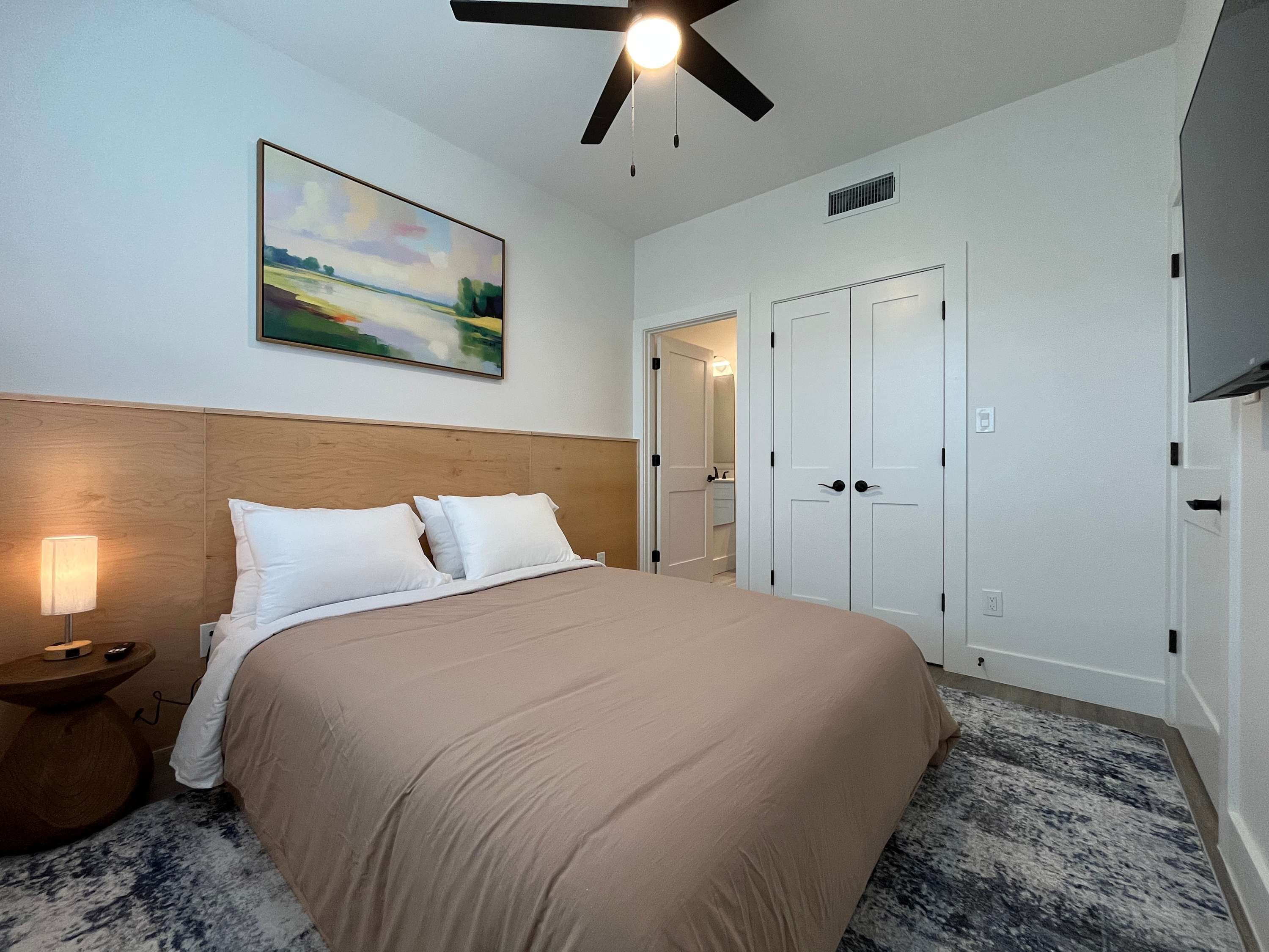 A hotel room with a bed, a nightstand, a ceiling fan, a painting, and a set of double doors.