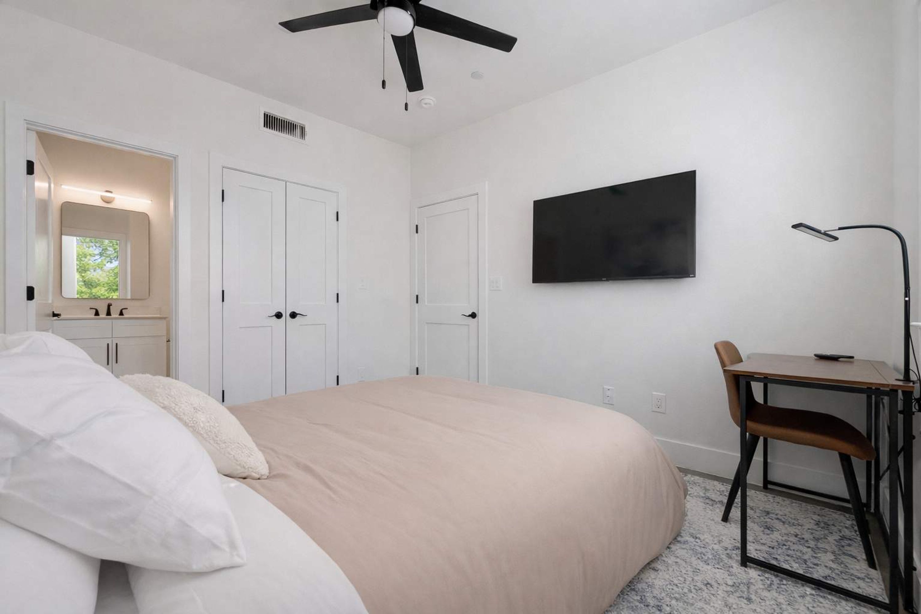 A bedroom with a bed, a desk, a chair, a ceiling fan, a TV, and a window.