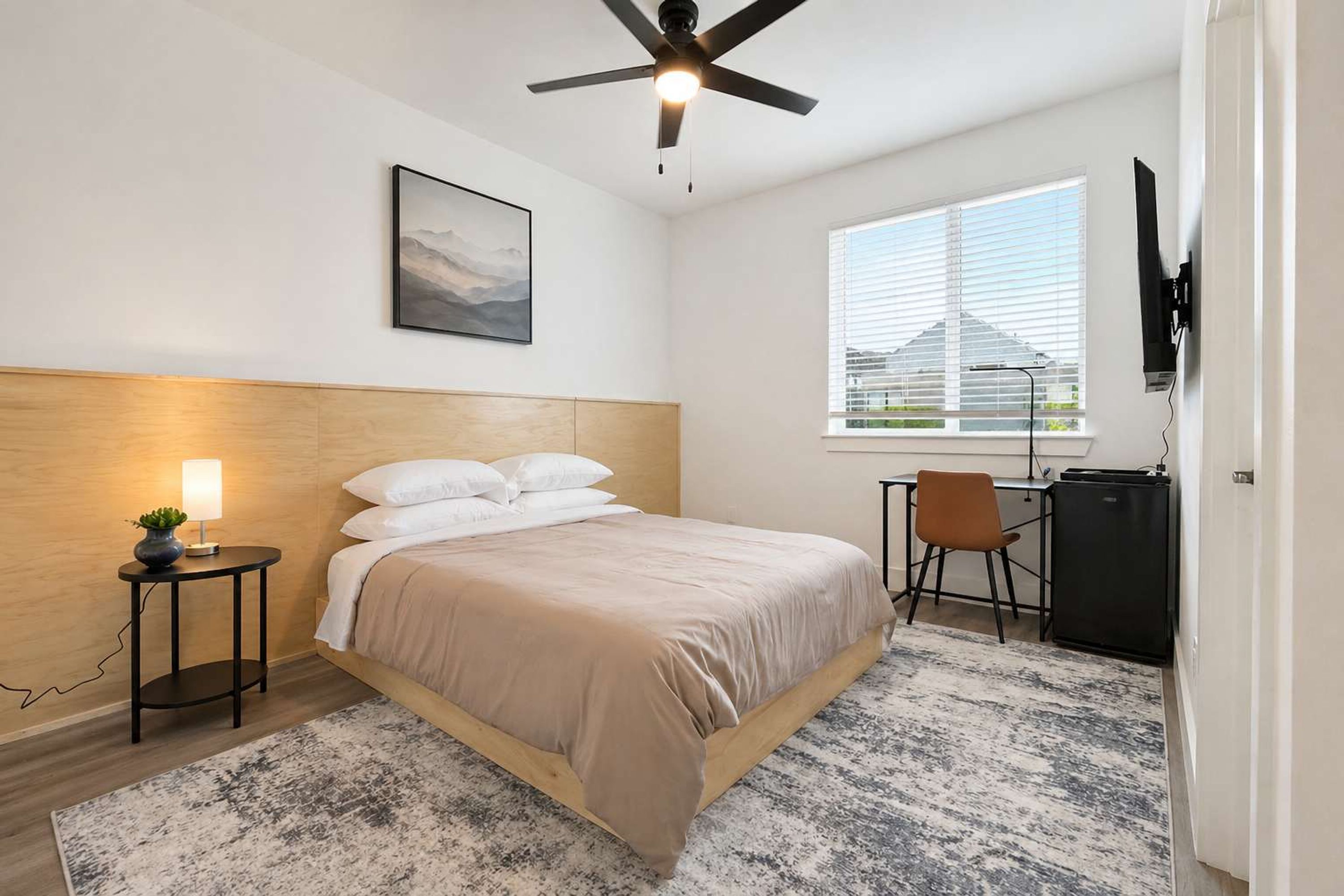 A bedroom with a bed, a desk, a chair, a ceiling fan, a window, and a wall-mounted TV.
