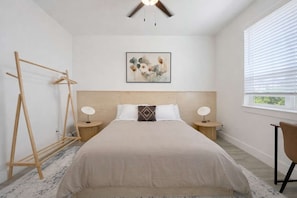 A bedroom with a bed, two bedside tables, a wooden headboard, a wall-mounted artwork, a window with blinds, and a wooden drying rack. - Bishop Arts 6BR Townhouse Sleeps 12 Close to DT (Dallas)