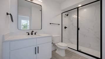 Un baño moderno con tocador blanco, un espejo grande, un inodoro y una ducha con mampara de vidrio.