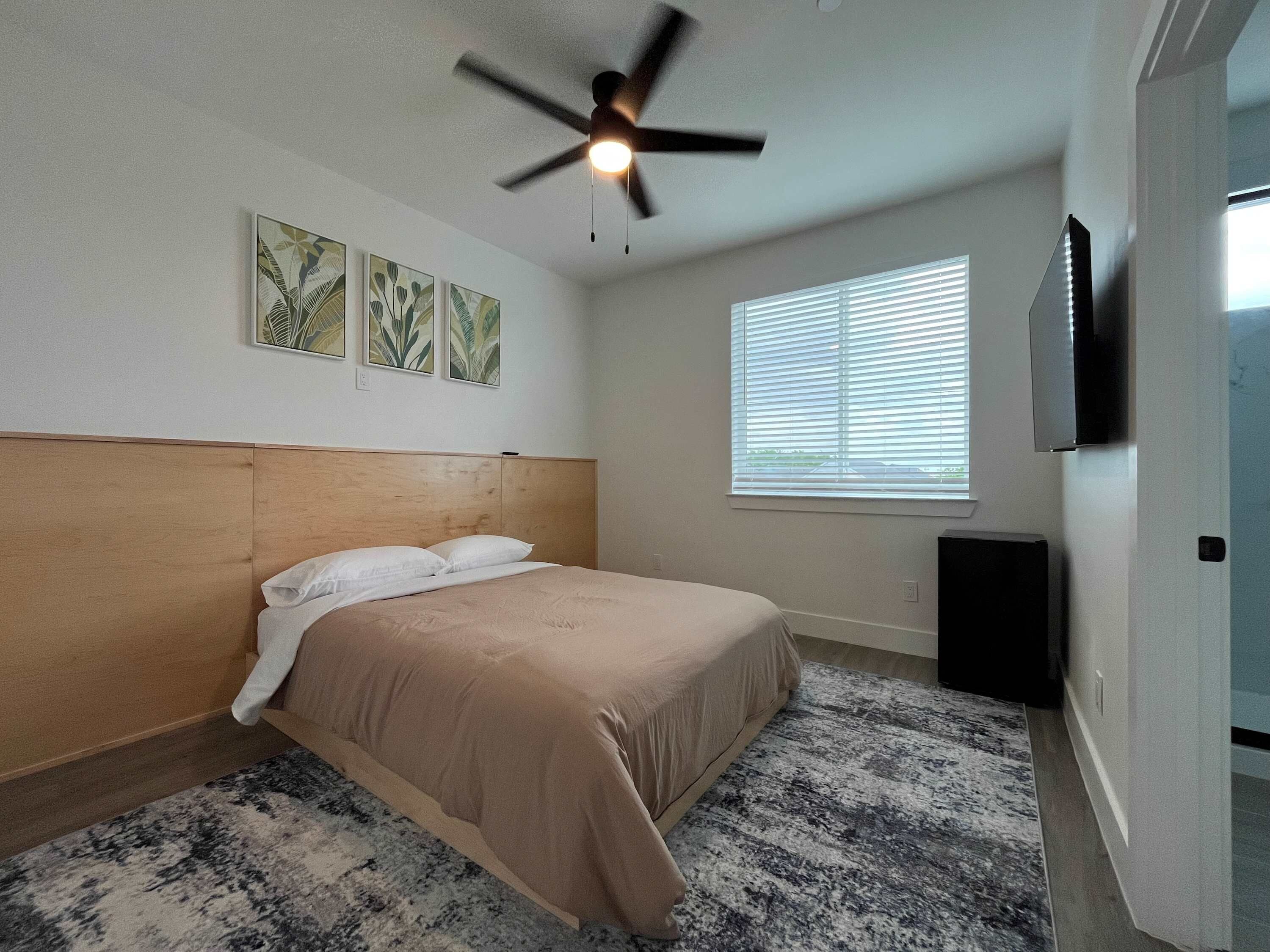 A bedroom with a bed, ceiling fan, wall-mounted TV, and window with blinds.