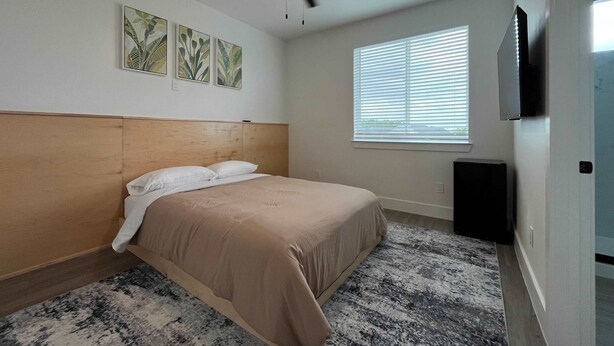 Un dormitorio con cama, ventilador de techo, televisor montado en la pared y ventana con persianas.