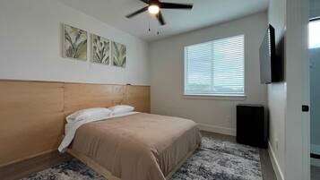 A bedroom with a bed, ceiling fan, wall-mounted TV, and window with blinds.