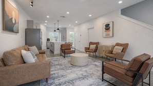 A modern living room with a sofa, chairs, and a coffee table. There is a kitchen in the background with cabinets and a refrigerator. - Bishop Arts 6BR Townhouse Sleeps 12 Close to DT (Dallas)