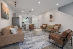 A modern living room with a sofa, chairs, and a coffee table. There is a kitchen in the background with cabinets and a refrigerator. - Bishop Arts 6BR Townhouse Sleeps 12 Close to DT (Dallas)