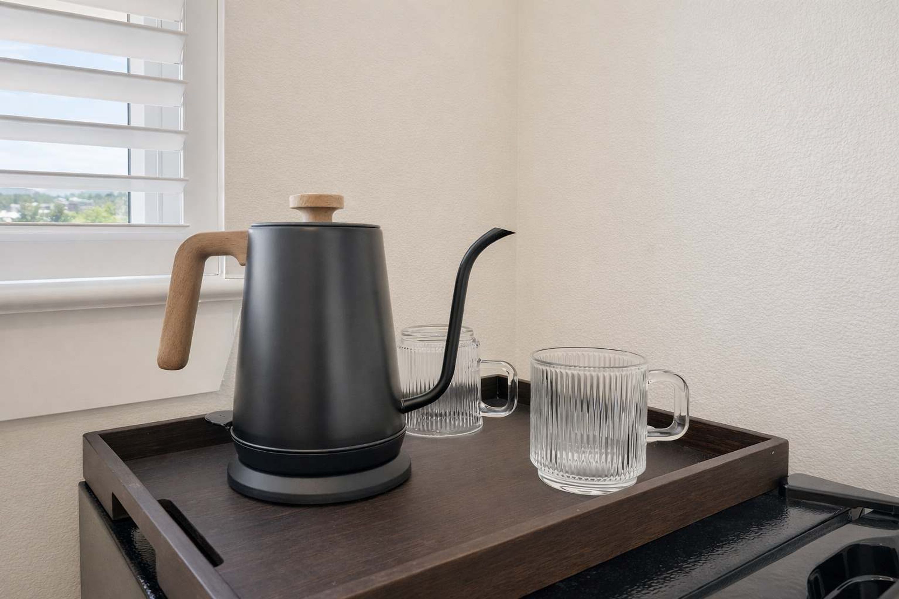 A modern kitchen area with a stainless steel kettle on a wooden tray, two clear glass mugs, and a window with blinds.