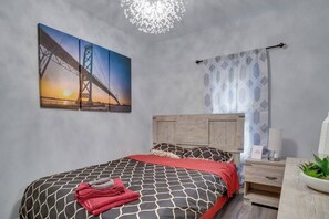 A bedroom with a bed, a nightstand, a lamp, a potted plant, and a painting of a bridge. - Modern & Private 1-Bedroom Apartment (Windsor)