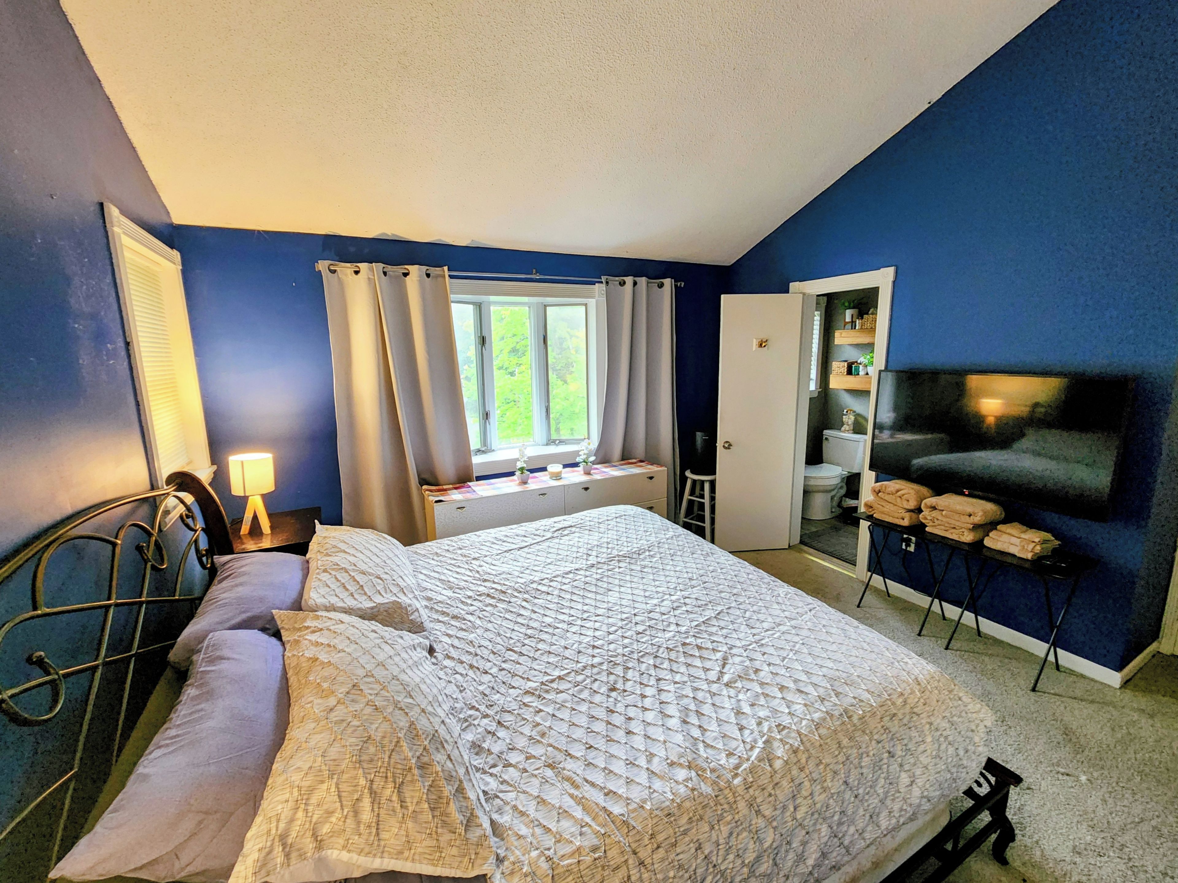 A bedroom with a bed, a nightstand, a lamp, a window with curtains, a television, and a bathroom door.