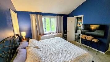A bedroom with a bed, a nightstand, a lamp, a window with curtains, a television, and a bathroom door.